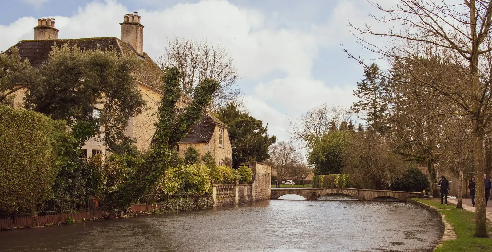 Couple on a low stone bridge over a stream in a Cotswold village of golden limestone cottages